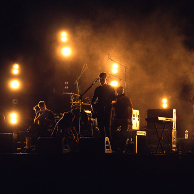 A picture of a band playing on stage on the dark with light behind them.